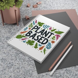 May include: A white spiral-bound notebook featuring the text "PLANT BASED" in a bold, dark font, surrounded by colourful botanical illustrations. Two pencils are positioned next to the notebook.