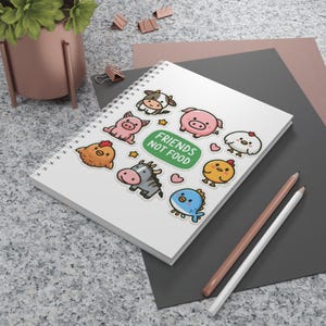 May include: A white spiral-bound notebook with cartoon animal stickers, such as a cow, pig, and chicken. The stickers surround a green rectangle with the text "FRIENDS NOT FOOD". Two pencils are on the right side of the notebook.