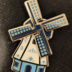 Op de afbeelding: Een gouden emaille pin met een blauwe en witte Nederlandse windmolen met een traditioneel ontwerp. De windmolen heeft blauwe wieken met witte vierkanten en een witte basis met blauwe en witte bloemmotieven.