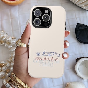 May include: A beige phone case with a car graphic and text that reads "Future Mrs. Boss Going to the Chapel!" is surrounded by accessories. These include a pearl clutch, sunglasses, jewellery, grapefruit halves, and seashells.