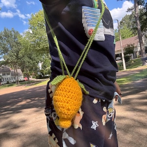 May include: A crocheted lemon-shaped pouch in bright yellow with green leaves, suspended by a green cord. The handmade accessory is worn around the neck. The pouch is a unique, whimsical item.