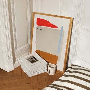 May include: A framed abstract painting with a red, orange, blue, and gray color scheme. The painting is leaning against a white wall next to a stack of magazines labeled 'ARK JOURNAL'. A white ceramic mug is in the foreground.