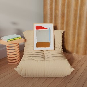 May include: A beige, tufted, oversized beanbag chair with a framed abstract painting on top. The painting has a red, blue, and brown color scheme. The chair is in front of a wood-paneled wall. A small, orange, spring-loaded stool with three books on top is to the left of the chair.