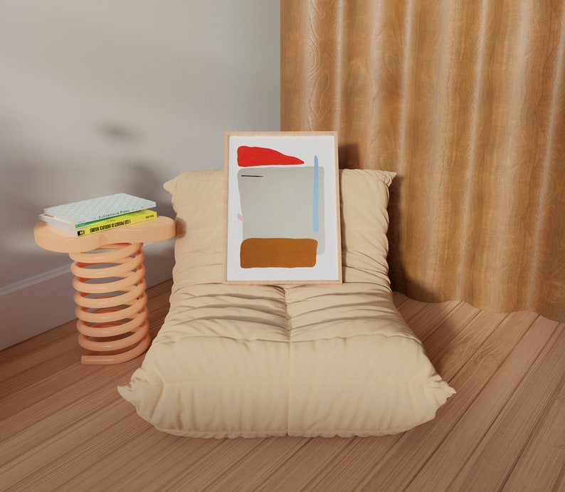 May include: A beige, tufted, oversized beanbag chair with a framed abstract painting on top. The painting has a red, blue, and brown color scheme. The chair is in front of a wood-paneled wall. A small, orange, spring-loaded stool with three books on top is to the left of the chair.