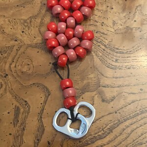 May include: A keychain featuring a heart-shaped charm constructed from alternating red and dusty pink plastic pony beads. A black elastic cord connects the heart to a vertical strand of three beads and two silver-colored aluminum soda can tabs.