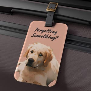 May include: A luggage tag with a peach background featuring a Golden Retriever illustration. The tag has the text "Forgetting Something?" in black script. The tag is attached to a black suitcase with a black strap and gold buckle.