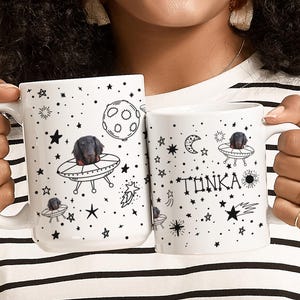May include: Two white ceramic mugs with black illustrations of a dog in a UFO, stars, planets, and the word "TONKA". The mugs have a handle and are held by a person wearing a striped shirt.