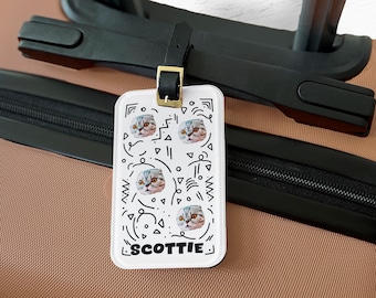 May include: A white luggage tag with a black leather strap and gold buckle. The tag features a repeating pattern of cat faces and abstract black shapes. The name "SCOTTIE" is printed in black at the bottom.