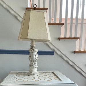 Könnte beinhalten: Ein weißer Beistelltisch mit goldenen Akzenten und einer dekorativen Cherub-Lampe mit einem weißen Lampenschirm. Der Tisch hat eine weiße Tischplatte mit einem goldenen geometrischen Muster.