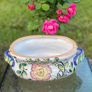 May include: A white ceramic serving bowl with a floral design. The bowl has a light brown rim and blue handles with yellow and green accents. Pink flowers and green leaves decorate the exterior. The bowl is approximately 30 cm in diameter.