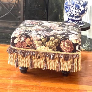 Puede incluir: Un reposapiés cuadrado con tapicería de tela marrón oscuro con un patrón floral multicolor en tonos rosa, crema y verde. La base está adornada con un fleco de cuerda grueso de color beige y descansa sobre cuatro patas pequeñas de color marrón oscuro.