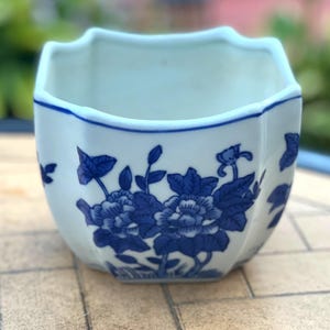 May include: A white ceramic planter with a scalloped rim and a blue floral pattern. The vessel features a repeating design of detailed blue flowers and leaves painted on the exterior surface, with a smooth white interior and a wide opening.