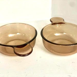 May include: Two amber-colored glass bowls with dark brown rims and handles. One bowl has a rounded handle, and the other has a flat, oval handle. The bowls are empty and sit on a white surface.