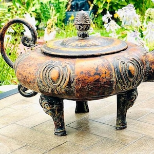 May include: A decorative, antique-style teapot with a distressed, brown and black finish. The teapot has a round body with intricate details and three legs. It is made of metal and has a spout and a lid.
