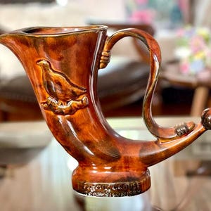 Könnte beinhalten: Ein braunes Trinkhorn aus Keramik mit einem Vogelrelief. Das hornförmige Gefäß hat einen gebogenen Griff und ein abgerundetes Ende. Auf der Basis steht "Famous Grouse Scotch Whisky."