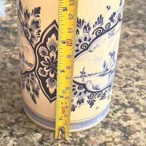 May include: A cylindrical ceramic vase with a cream-colored base and blue floral and windmill designs. The vase is approximately 5 inches tall, with a yellow measuring tape next to it.