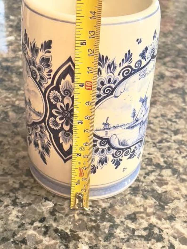 May include: A cylindrical ceramic vase with a cream-colored base and blue floral and windmill designs. The vase is approximately 5 inches tall, with a yellow measuring tape next to it.
