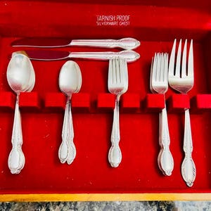 Puede incluir: Un estuche de madera para cubiertos con interior de terciopelo rojo que contiene cubiertos plateados, incluyendo cuchillos, cucharas y tenedores. La tapa interior muestra el texto TARNISH PROOF SILVERWARE CHEST. Los cubiertos tienen mangos decorativos.