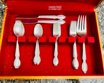 May include: A wooden silverware chest with a red velvet interior containing silver-toned flatware including knives, spoons, and forks. The interior lid features the text TARNISH PROOF SILVERWARE CHEST. The utensils have decorative handles.