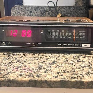 May include: Vintage Sears dual alarm clock radio with a wood-grain finish. The digital display shows 12:00 PM in red. Features an AM/FM radio dial and the text "DUAL ALARM BATTERY BACK-UP".