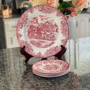 May include: A set of vintage red and white plates with a detailed transferware design. The plates feature a rural scene with a horse-drawn carriage and floral border. A large plate is displayed on a wooden stand, with a stack of smaller plates in front.
