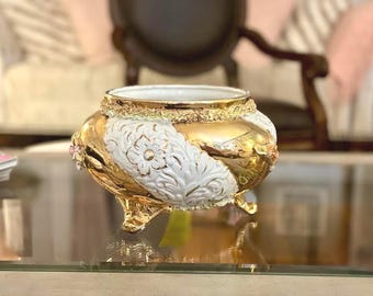 Puede incluir: Un tazón de cerámica blanca con detalles dorados y diseños florales. El tazón tiene tres patas doradas y está decorado con flores rosas.