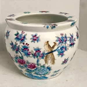 Könnte beinhalten: Weißer Keramik-Blumentopf mit breiter Öffnung und rundem Körper. Der Topf ist mit blauen, rosa und braunen Blumen- und Vogelmotiven verziert. Der Rand weist ein sich wiederholendes Blumenmuster auf.