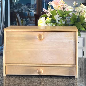 May include: A light-colored wooden bread box featuring a rectangular design with a drop-down front door and a small storage drawer at the bottom. Both the door and the drawer are equipped with a single, centered, round wooden knob for opening.