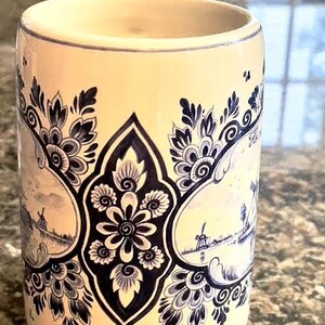 May include: Cream-colored ceramic mug with blue floral and landscape designs. The mug features a central floral motif and framed landscape scenes with windmills. The rim and base have blue accents, creating a classic, decorative look.