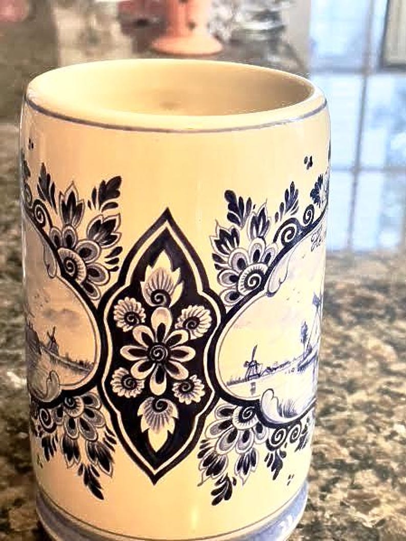 May include: Cream-colored ceramic mug with blue floral and landscape designs. The mug features a central floral motif and framed landscape scenes with windmills. The rim and base have blue accents, creating a classic, decorative look.