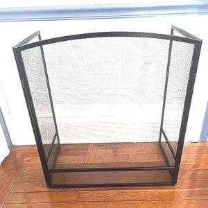 Può includere: Un parascintille in metallo nero con un centro a rete. Lo schermo ha una cornice rettangolare con una parte superiore curva ed è progettato per proteggere da scintille e braci. È posto su un pavimento di legno.