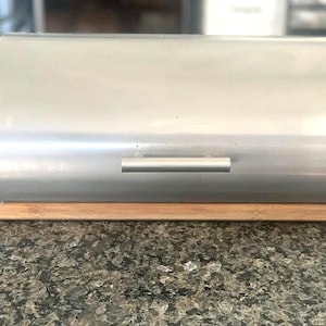 May include: A stainless steel bread box with a curved top and a wooden base. The box has a silver handle and is designed to store bread and baked goods. The bread box is sitting on a granite countertop.
