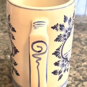 May include: Cream-colored ceramic mug with a blue floral design and a matching handle. The mug features a blue spiral design on the handle and a blue rim. The mug is a classic design for enjoying hot beverages.