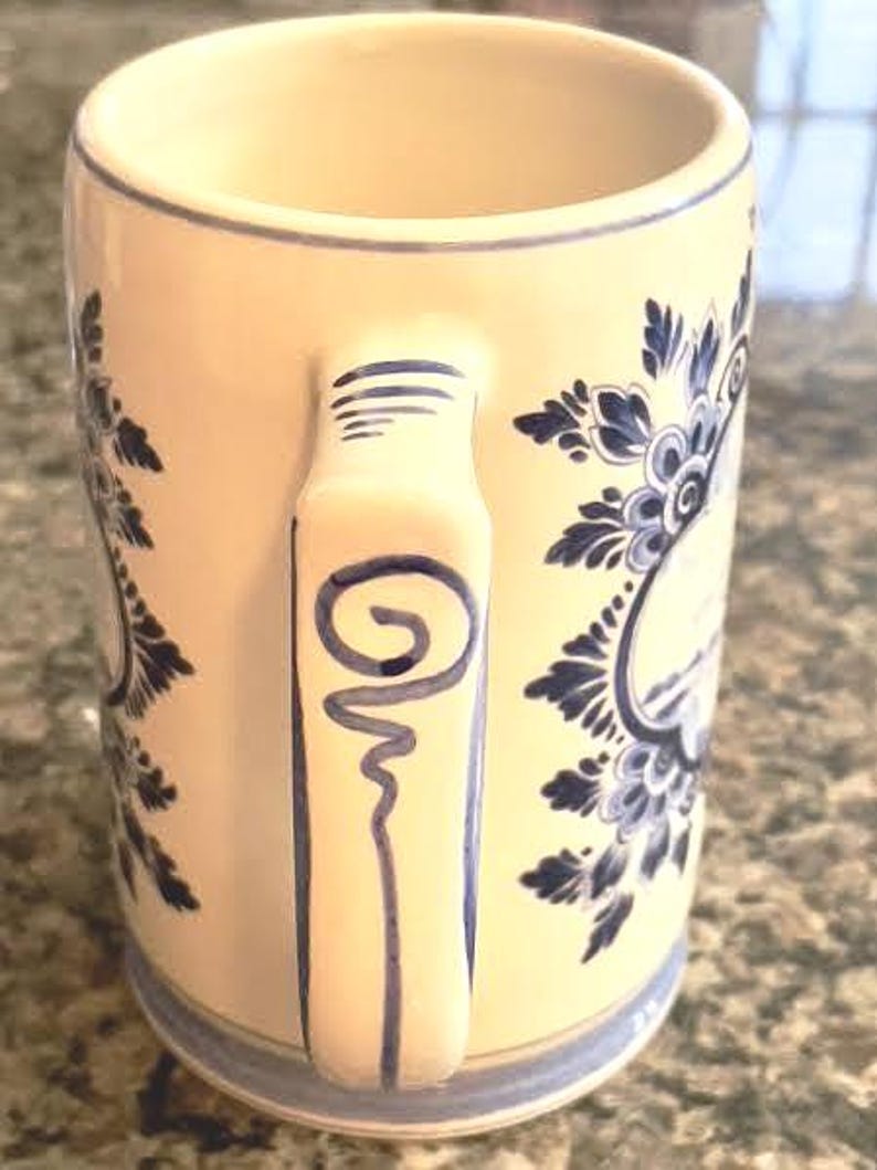 May include: Cream-colored ceramic mug with a blue floral design and a matching handle. The mug features a blue spiral design on the handle and a blue rim. The mug is a classic design for enjoying hot beverages.