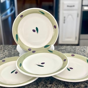 Pode incluir: Quatro pratos de cerâmica branca com uma borda verde e azeitona. Cada prato apresenta um desenho de ramo de oliveira pintado com folhas verdes e azeitonas roxas. Os pratos estão dispostos em uma bancada salpicada.