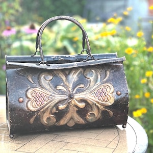 May include: A dark brown metal handbag with a distressed finish and a prominent embossed floral pattern in a lighter tan colour. The bag features a single arched handle, a hinged top opening, and four small spherical feet at the base.