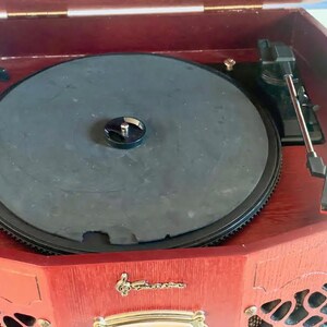 May include: A classic record player in a dark wood cabinet. The black turntable is visible, along with the tonearm and needle. The front panel features decorative elements and a gold-coloured emblem.