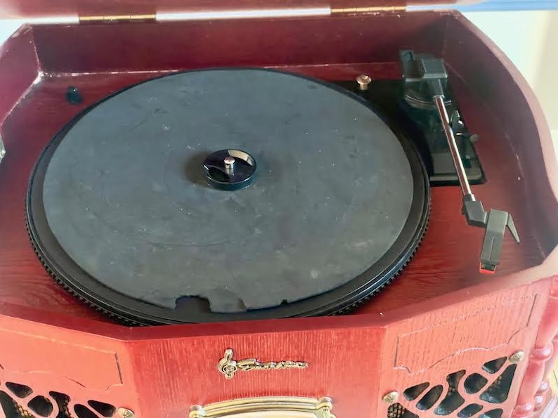 May include: A classic record player in a dark wood cabinet. The black turntable is visible, along with the tonearm and needle. The front panel features decorative elements and a gold-coloured emblem.
