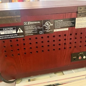 May include: A vintage Emerson MR1117T radio with a dark wood finish. The back of the radio features a series of small circular vents, a line out port, and various warning labels. The radio was manufactured in August 2007.