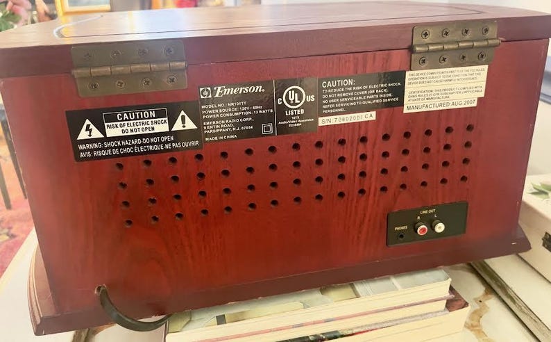 May include: A vintage Emerson MR1117T radio with a dark wood finish. The back of the radio features a series of small circular vents, a line out port, and various warning labels. The radio was manufactured in August 2007.