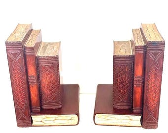 Puede incluir: Un par de sujetalibros, cada uno diseñado para parecer una pila de tres libros verticales sobre una base de libro horizontal. Los sujetalibros tienen un acabado marrón rojizo oscuro con patrones entrecruzados y bordes de página color crema.