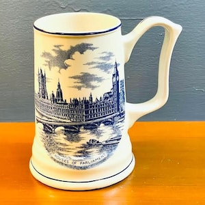 Può includere: Una tazza in ceramica bianca con un'illustrazione blu e bianca delle Case del Parlamento a Londra. La tazza ha un manico e un bordo blu.