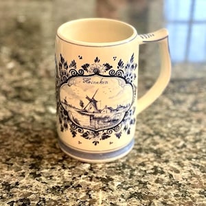 May include: Cream-colored ceramic beer stein with a blue and white windmill design. The stein has a handle and a decorative border. The word "Heineken" is written on the front of the stein.