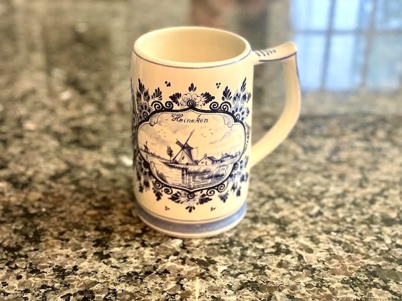 May include: Cream-colored ceramic beer stein with a blue and white windmill design. The stein has a handle and a decorative border. The word "Heineken" is written on the front of the stein.