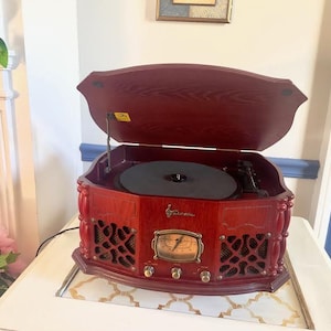 May include: A vintage-style, red wooden record player with a hinged lid. The player features a black turntable, a gold-toned dial, and decorative speaker grilles. It sits on a white surface.