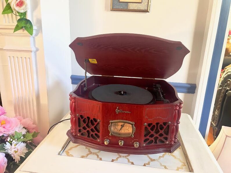 May include: A vintage-style, red wooden record player with a hinged lid. The player features a black turntable, a gold-toned dial, and decorative speaker grilles. It sits on a white surface.