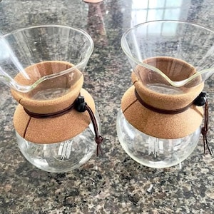 May include: Two clear glass coffee makers with a cork band around the middle and a glass top. The coffee makers have a brown leather string and a black bead. The coffee makers are on a speckled gray surface.