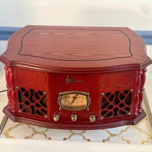 May include: A red wooden radio with a dark top and decorative detailing. The radio has a gold-coloured dial and knobs, and speaker grilles. The design is reminiscent of classic audio equipment.