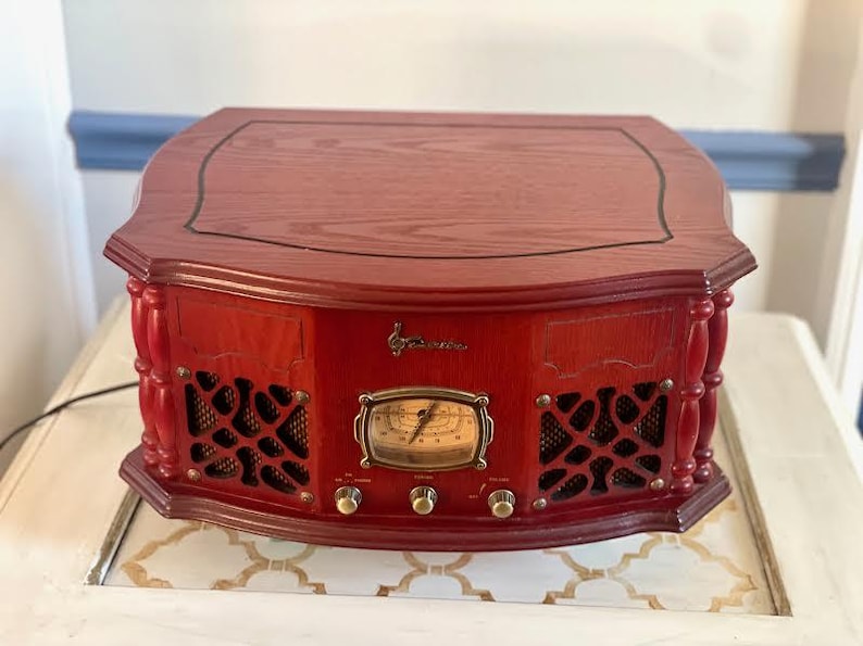May include: A red wooden radio with a dark top and decorative detailing. The radio has a gold-coloured dial and knobs, and speaker grilles. The design is reminiscent of classic audio equipment.