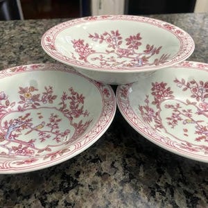 Puede incluir: Tres cuencos de porcelana blanca con un diseño floral y de pájaros en rosa. Los cuencos tienen un borde festoneado con un borde rosa. El interior de los cuencos presenta una ilustración detallada de flores, ramas y pájaros. Los cuencos están colocados sobre una encimera moteada.